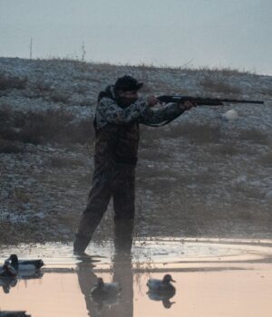 Man aiming rifle near pond with ducks during hunting session in misty dawn.