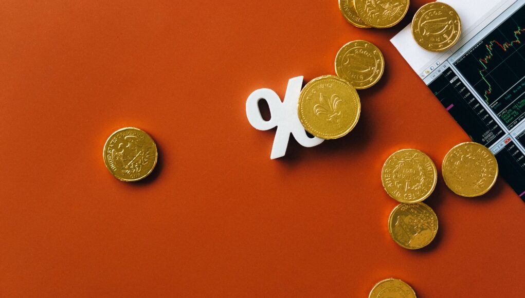 pexels-photo-6120252 Gold coins scattered with a stock market graph and a percentage symbol on an orange background.