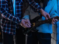GTA VI Is Not Just Getting a Soundtrack. It Is Getting a Music Business Two young adults playing an electric guitar together in a cozy indoor setting, showcasing teamwork and learning.