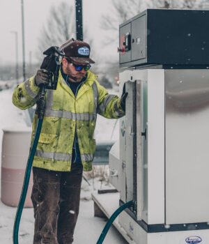 A man wearing a jacket refuels a truck at a gas station during winter with snow falling.