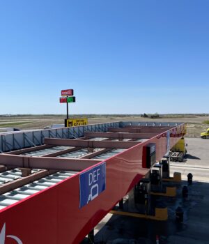 pexels-photo-12249509 Aerial view of a truck stop with gas station and parking lot under a clear blue sky.