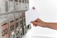 California’s Ballot Lawsuit Is a Test of Who Decides Voter Eligibility Close-up of a hand inserting a stamped envelope into a mailbox, symbolizing voting.