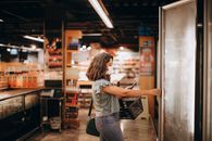 The Supermarket Refrigeration Hack Is Really a Remote-Access Failure Woman wearing mask shops in grocery store, reaching for freezer door, holding basket.