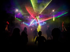 The Internet’s Most Embarrassing Memories Are Becoming Nightlife Silhouettes dance amid colorful laser lights in a vibrant nightclub setting.