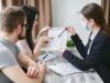 Open Enrollment Is Here. Families May Keep Insurance and Lose Their Children’s Hospital Three adults discuss a home insurance policy at a meeting table indoors.